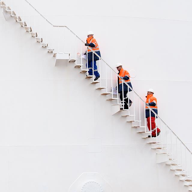 three men in orange and wearing hardhats climbing storage tank stairs